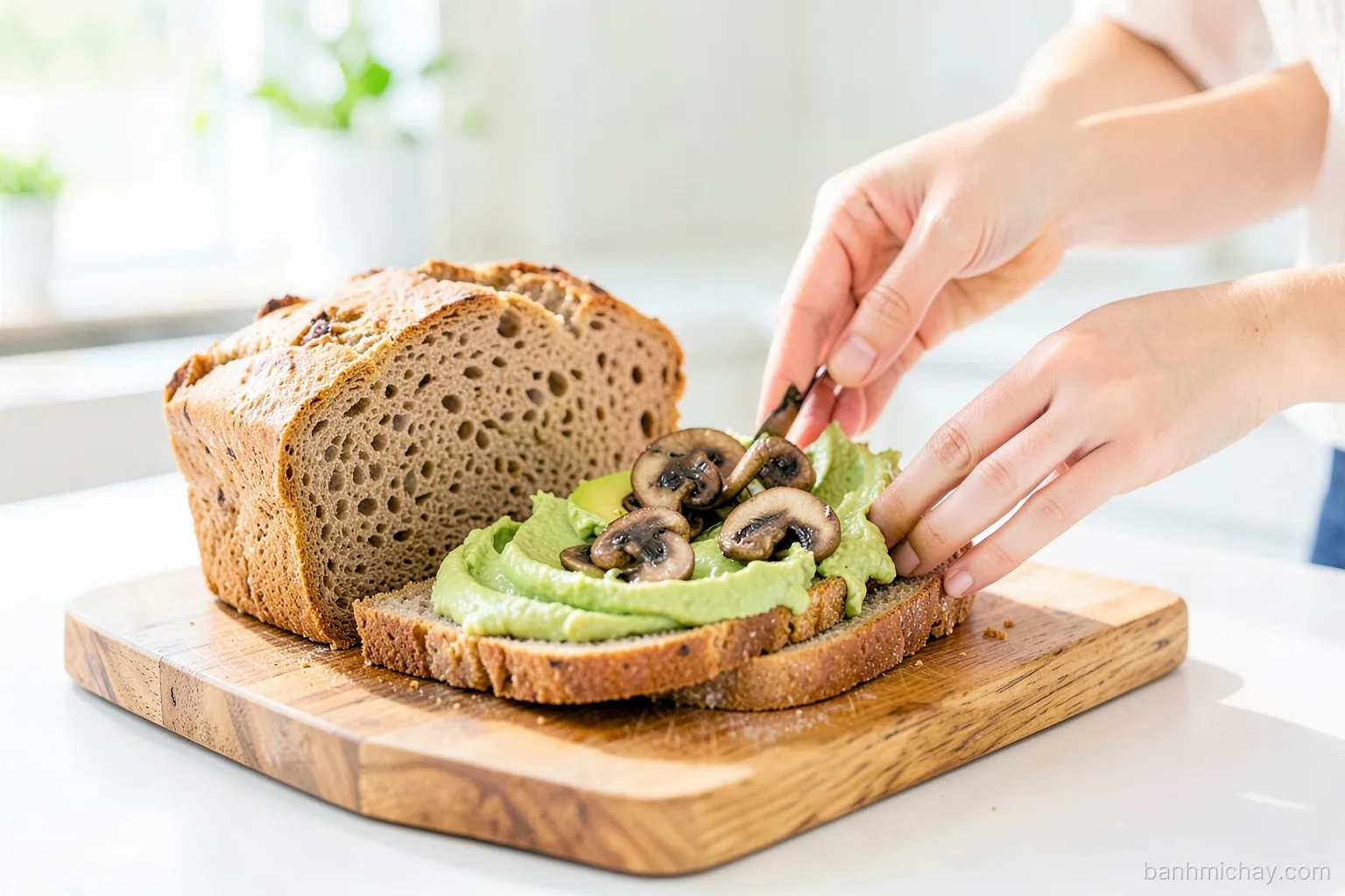 Sliced whole wheat bread loaf on a wooden board with hummus, avocado and sauteed mushrooms being spread on slices, Vietnamese woman hands, bright morning kitchen, close-up