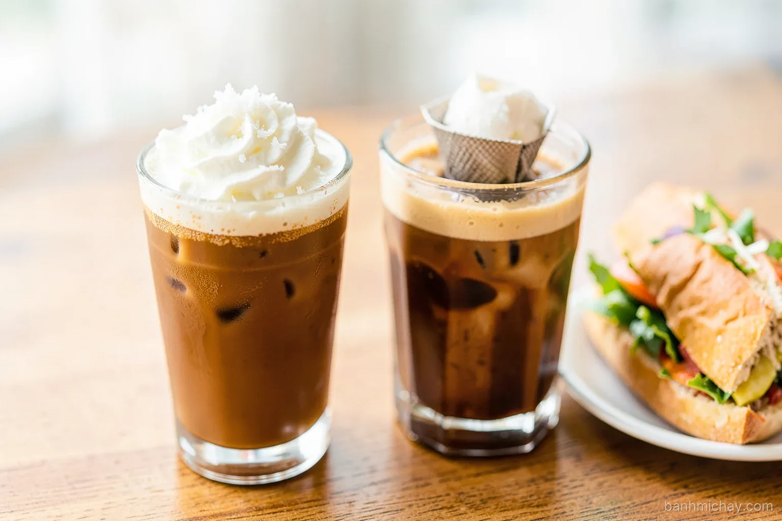 Close-up of two glasses of Vietnamese iced coffee on a wooden cafe table, one topped with whipped coconut salt cream, phin filter dripping beside a vegetarian banh mi, soft light