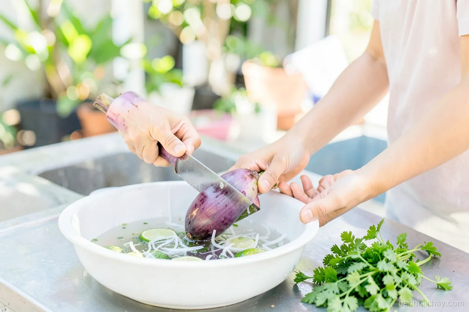 Vietnamese woman thinly slicing a purple banana blossom over a basin of lime water in an outdoor kitchen, white shreds floating, Vietnamese coriander nearby, close-up side angle, morning light