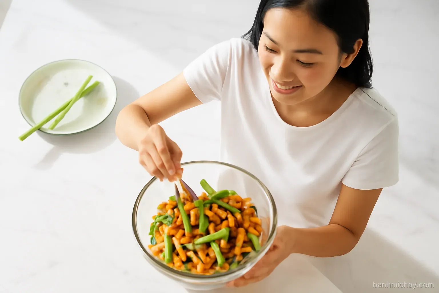 Overhead shot of a young Vietnamese woman tossing lotus stems, carrot ribbons and fried tofu in a glass bowl on a white marble counter, soft morning light, airy editorial style