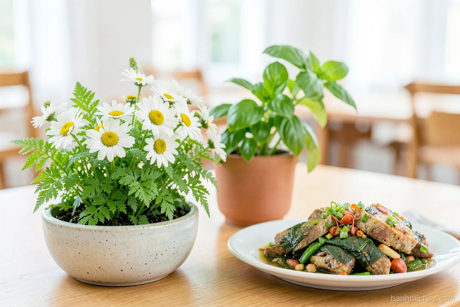 Low ceramic bowl of white daisies and fern leaves beside potted Thai basil and a vegan banh mi platter, Vietnamese dining room, soft daylight, three-quarter angle