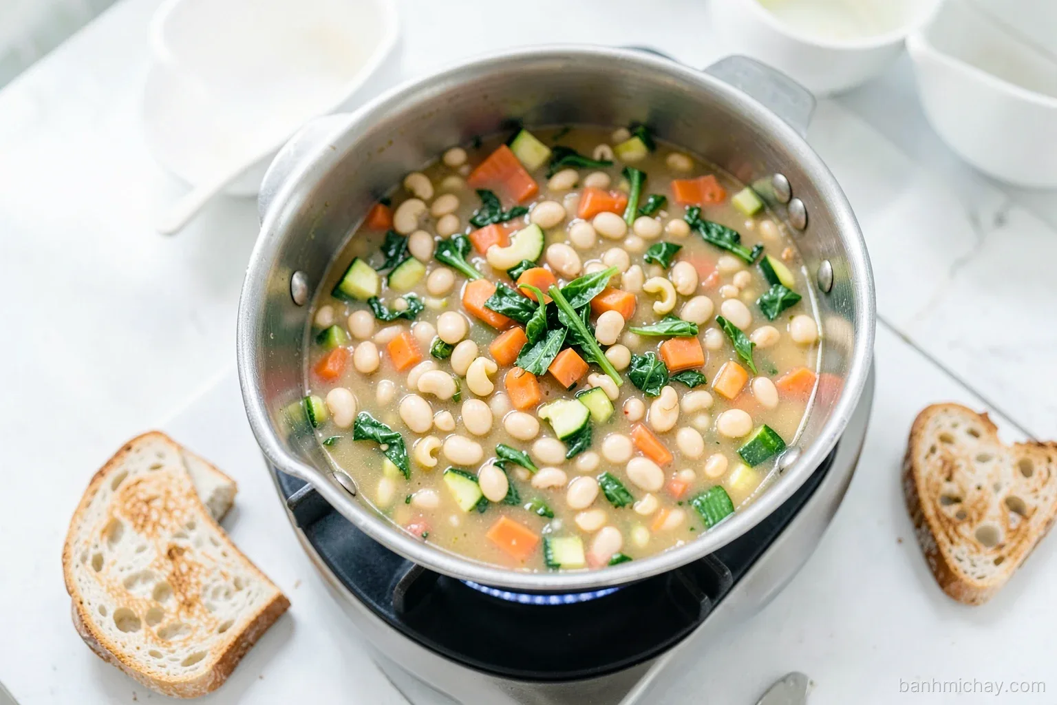 Large pot of minestrone with white beans, carrot, zucchini, small pasta and greens on a gas stove in a Vietnamese apartment kitchen, toasted baguette slices nearby, top-down view