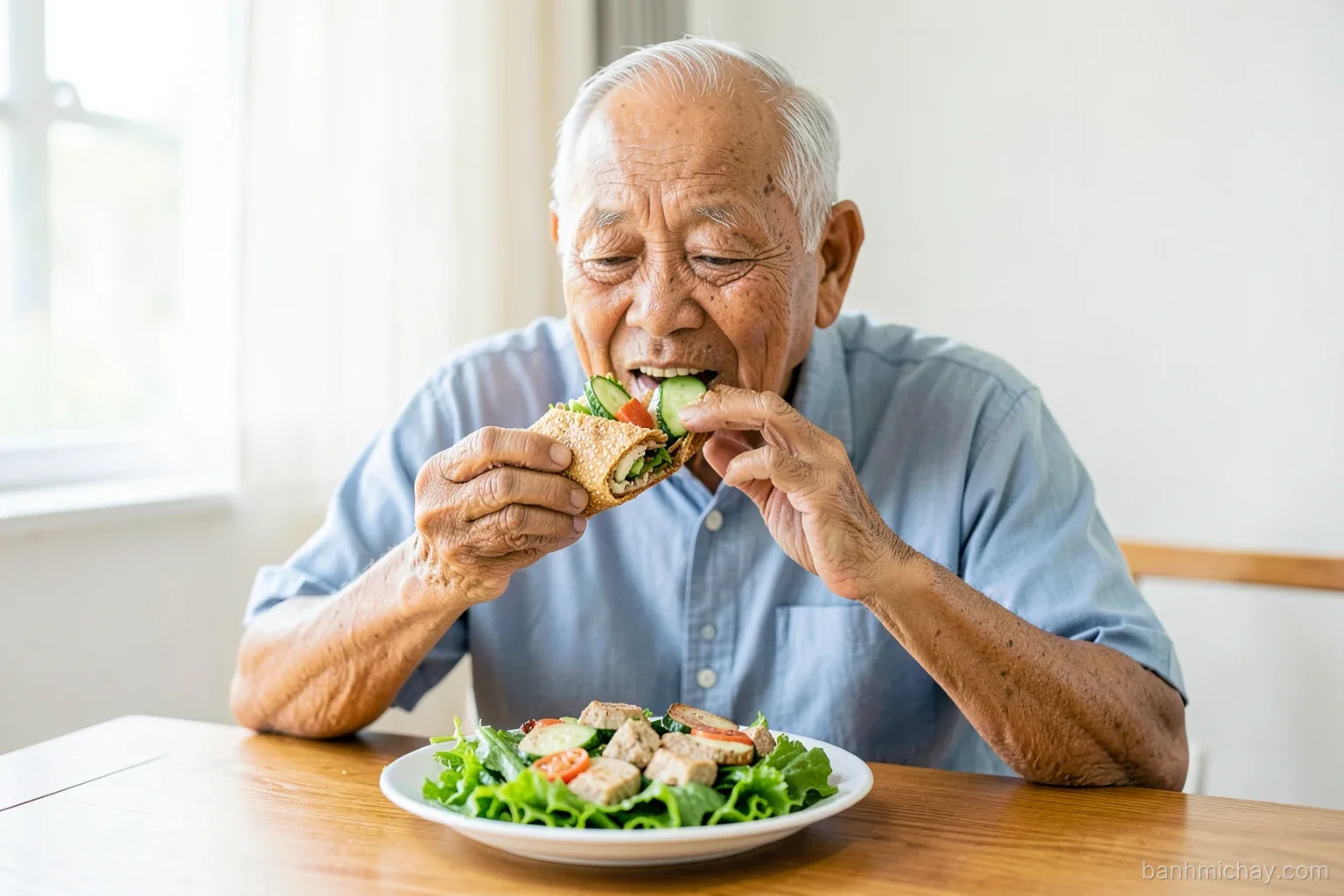 Elderly Vietnamese man at a wooden home table eating half a whole wheat banh mi with tofu and cucumber, plate of fresh greens, soft window light, eye level