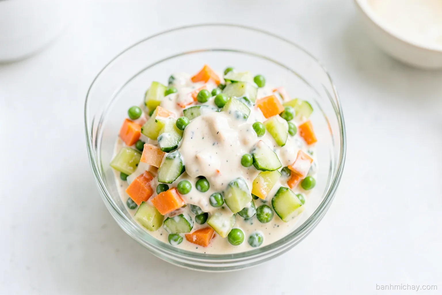 Top-down view of a glass bowl of vegan Russian potato salad with diced carrots, green peas and pickled cucumber in creamy soy mayo, Vietnamese kitchen counter, soft morning light