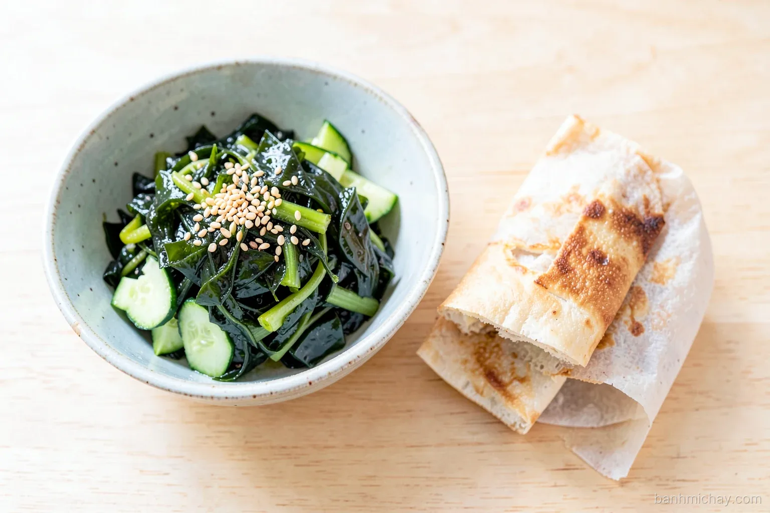 Top-down view of a ceramic bowl of glossy wakame seaweed salad with cucumber and toasted sesame beside a halved tofu banh mi on a light wooden table, soft daylight