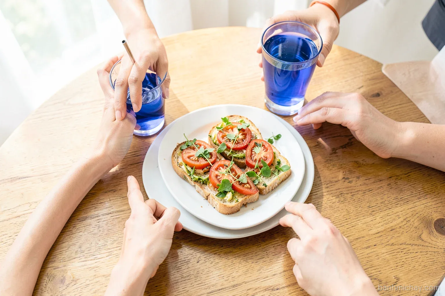 Overhead view of a wooden table with mini vegan banh mi, crostini topped with tomato and herbs, glasses of blue butterfly pea tea, Vietnamese women hands reaching, afternoon window light